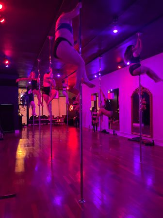 Energetic pole fitness class in a pink-purple lit studio — participants practicing spins and climbs on chrome poles beside mirrors over a polished hardwood floor.