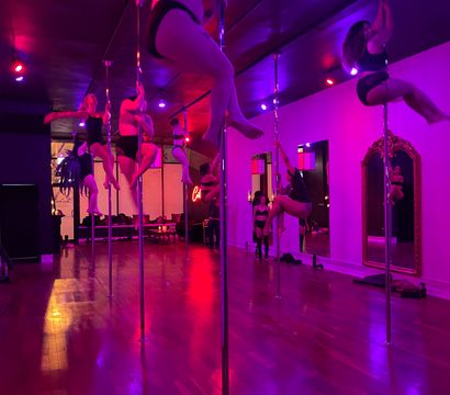 Energetic pole fitness class in a pink-purple lit studio — participants practicing spins and climbs on chrome poles beside mirrors over a polished hardwood floor.