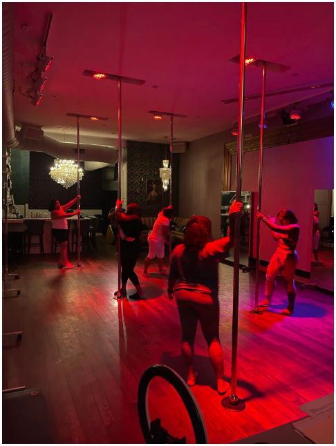 Group pole fitness class in a dim, red-lit dance studio with chrome poles, hardwood floors, mirrors and a chandelier.