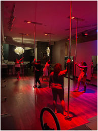 Group pole fitness class in a dim, red-lit dance studio with chrome poles, hardwood floors, mirrors and a chandelier.