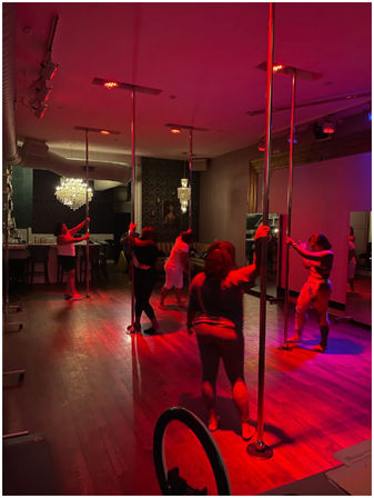 Group pole fitness class in a dim, red-lit dance studio with chrome poles, hardwood floors, mirrors and a chandelier.