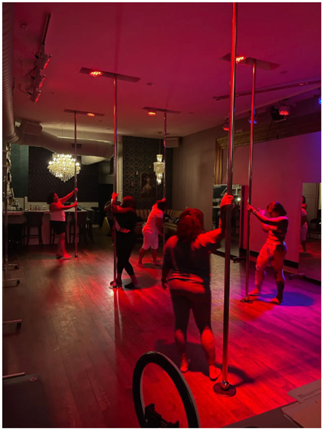 Group pole fitness class in a dim, red-lit dance studio with chrome poles, hardwood floors, mirrors and a chandelier.