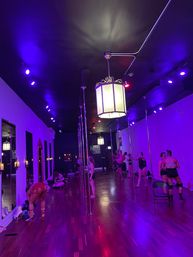 Pole fitness studio interior with purple LED lighting, hardwood floor, hanging lanterns, mirrored wall and students practicing on vertical poles.