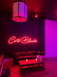Moody pink-purple lounge with a neon script wall sign, tufted velvet sofa, wooden coffee table with lit candles, vintage pendant light and hardwood floors.