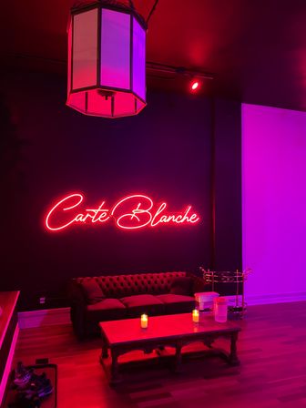 Moody pink-purple lounge with a neon script wall sign, tufted velvet sofa, wooden coffee table with lit candles, vintage pendant light and hardwood floors.