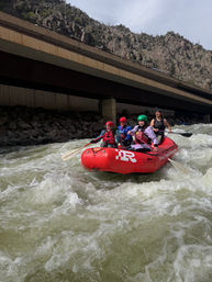 Four paddlers in a red inflatable navigating turbulent whitewater rapids under a highway bridge in a rocky mountain canyon