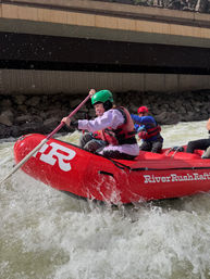 Whitewater rafting adventure: rafters in a red inflatable navigating choppy rapids under a concrete bridge, wearing helmets and life jackets as water splashes around them.