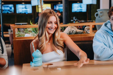 Smiling woman at an indoor bar lounge reaching to push a tabletop shuffleboard puck, blue frog-shaped novelty drink with striped straw on the wooden table, TVs and plants in the background.