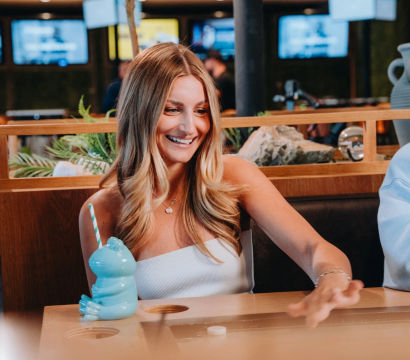 Smiling woman at an indoor bar lounge reaching to push a tabletop shuffleboard puck, blue frog-shaped novelty drink with striped straw on the wooden table, TVs and plants in the background.