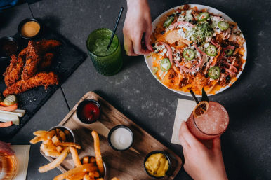 Top-down photo of a shared pub table with cheesy loaded nachos topped with jalapeños and pickled onions, crispy chicken tenders and fries with dipping sauces, and two colorful cocktails.