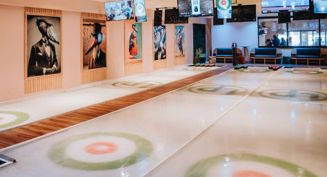 Bright indoor recreational curling rink with multiple lanes and painted target rings, wooden walkways, overhead TV screens, bench seating and playful framed bird portraits on the wall
