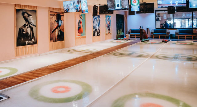 Bright indoor recreational curling rink with multiple lanes and painted target rings, wooden walkways, overhead TV screens, bench seating and playful framed bird portraits on the wall