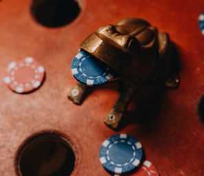 Close-up of a brass frog figurine holding a blue poker chip on a worn wooden game board with scattered red and blue chips