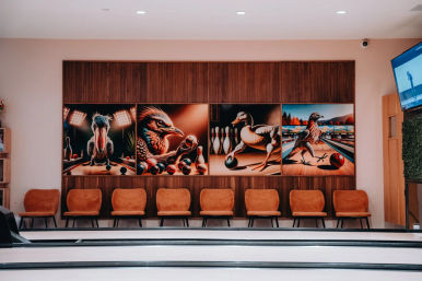 Bright bowling alley interior with multiple lanes, a row of orange mid-century chairs and a four-panel mural of birds playing with bowling balls and pins above the seating area.