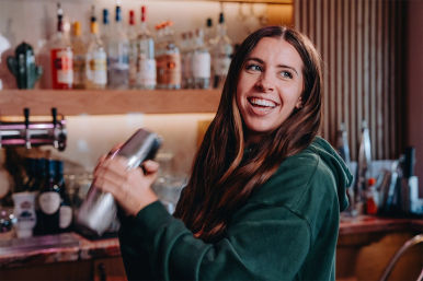 Smiling bartender energetically shaking a metal cocktail shaker behind a warm, wood-shelved bar with bottles and taps