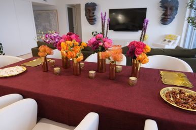 Open-plan modern dining table with burgundy tablecloth and gold vases holding vibrant orange, pink and purple flower arrangements — stylish indoor centerpiece for entertaining.