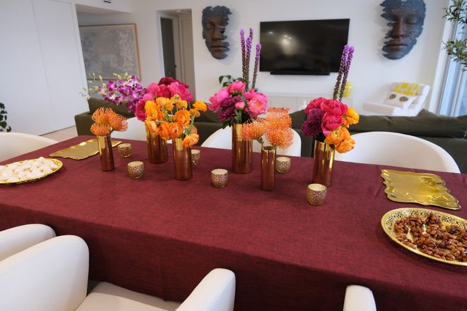 Open-plan modern dining table with burgundy tablecloth and gold vases holding vibrant orange, pink and purple flower arrangements — stylish indoor centerpiece for entertaining.