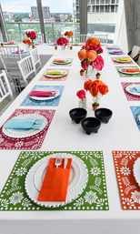Vibrant rooftop dining on a high-rise balcony: long white table with colorful patterned placemats, folded napkins, beaded white plates, bright orange and pink floral centerpieces and black bowls, city skyline visible through floor-to-ceiling windows.