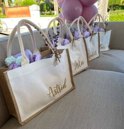 Sunlit outdoor patio scene with a row of jute-and-canvas tote gift bags tied with twine and lavender sprigs, filled with purple tissue and pom-pom favors, neatly lined on a beige outdoor sofa beneath pastel balloons for a garden party.