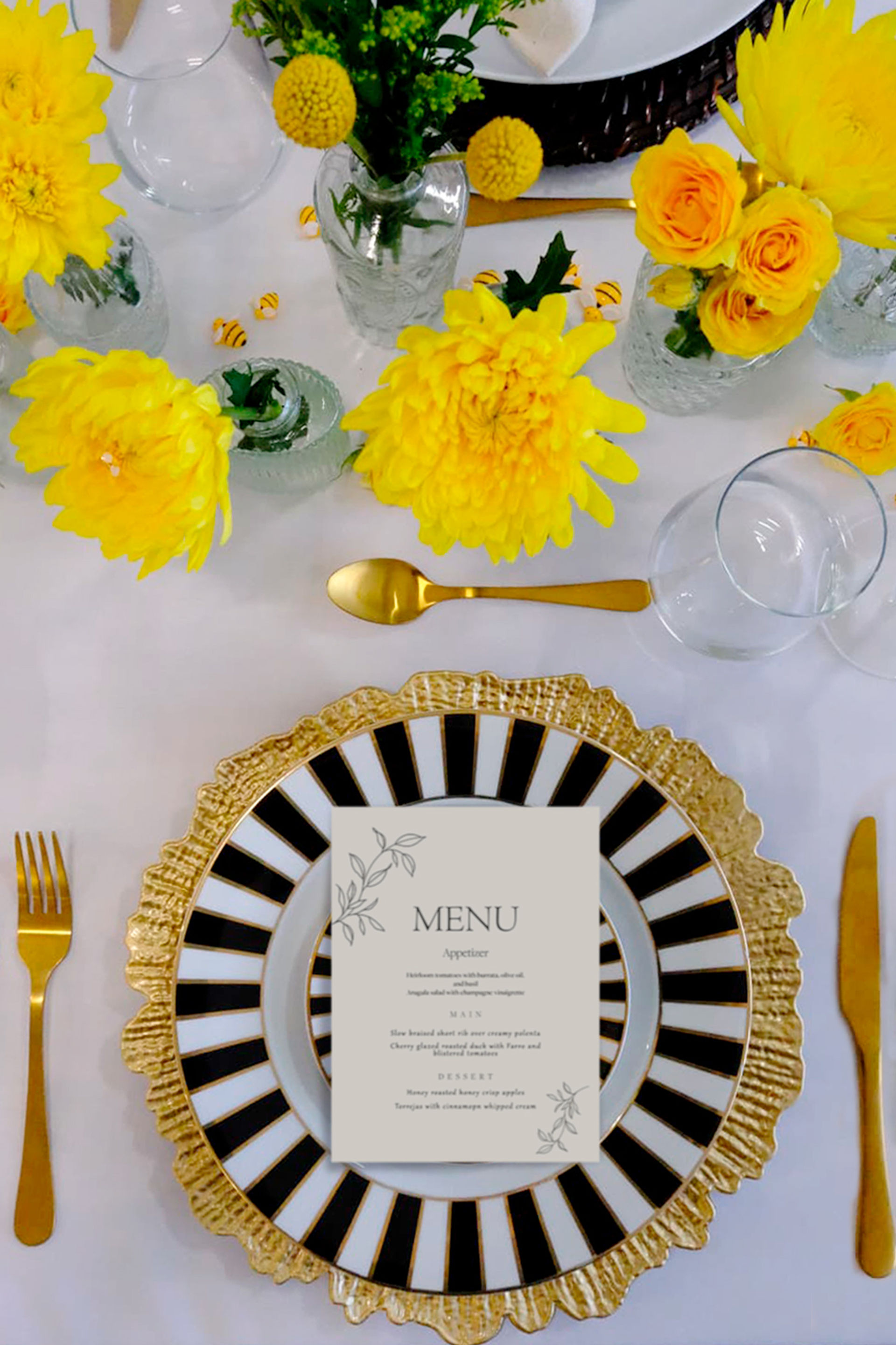 Cheerful yellow floral centerpiece in glass vases over a formal white tablescape featuring a black-and-white striped plate on a gold scalloped charger, gold flatware, wine glass, and a printed menu card.