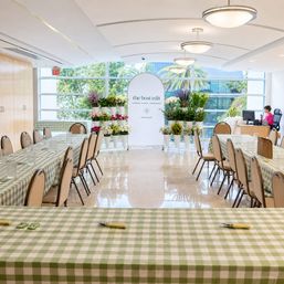 Bright indoor floral workshop and event space with green gingham-covered long tables, rows of chairs, white buckets of colorful flowers near large windows with palm trees outside, and a staff desk in the back.
