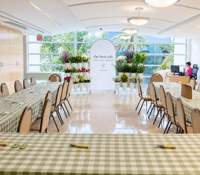 Bright indoor floral workshop and event space with green gingham-covered long tables, rows of chairs, white buckets of colorful flowers near large windows with palm trees outside, and a staff desk in the back.