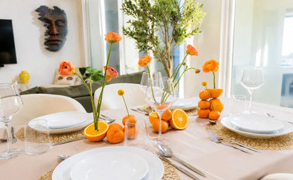 Sunlit modern dining table set with white plates and wine glasses, playful citrus centerpiece of halved oranges, stacked tangerines and orange ranunculus blooms on woven placemats in a bright living room