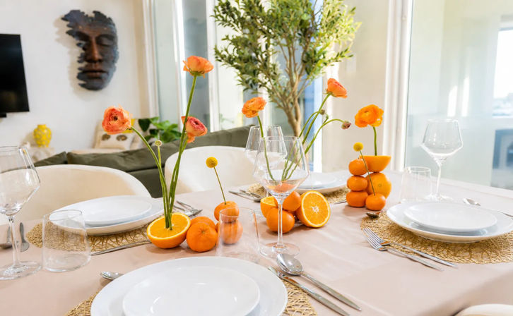 Sunlit modern dining table set with white plates and wine glasses, playful citrus centerpiece of halved oranges, stacked tangerines and orange ranunculus blooms on woven placemats in a bright living room