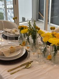 Sunny yellow floral tablescape with chrysanthemums and billy balls in clear glass vases, white plates on woven chargers, linen napkins with honeycomb napkin rings, gold flatware and assorted glassware on a cream tablecloth by a windowed balcony.