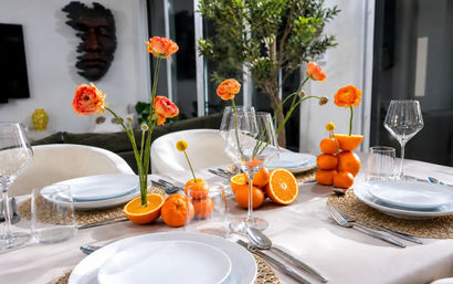 Stylish contemporary dining table in a bright interior, set with white plates, gold woven placemats and crystal wine glasses, featuring a fresh citrus centerpiece of stacked and halved oranges paired with orange ranunculus blooms.