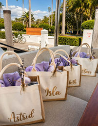 Cream canvas tote gift bags filled with lavender-colored goodies and dried lavender sprigs, lined up on a yacht bench at a sunny palm-tree marina dock.