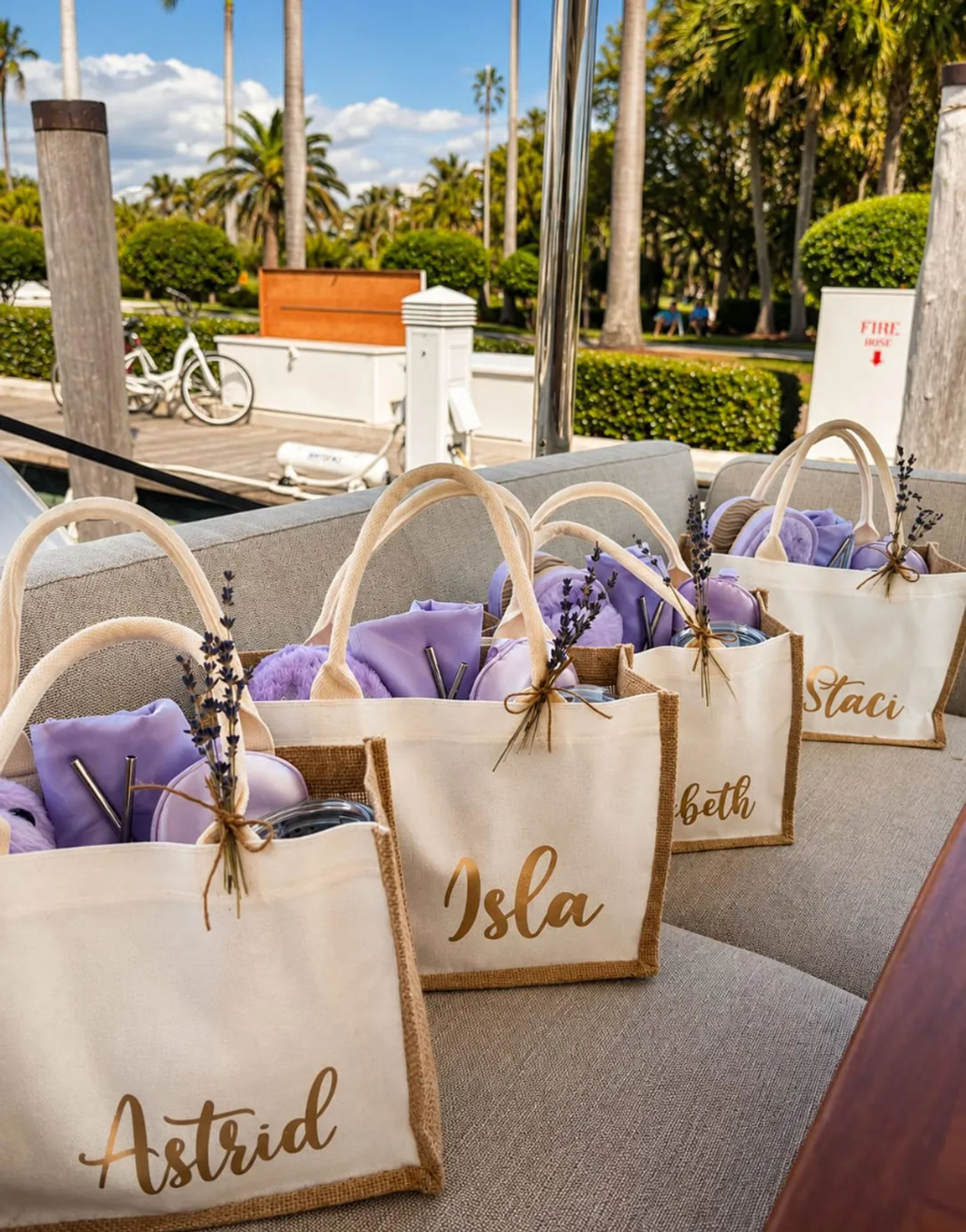 Cream canvas tote gift bags filled with lavender-colored goodies and dried lavender sprigs, lined up on a yacht bench at a sunny palm-tree marina dock.