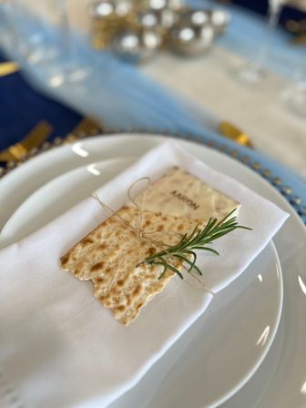 Chic place setting with stacked white plates and a folded napkin topped by a twine-tied flat cracker and fresh rosemary sprig, soft blue and gold table decor blurred in the background.