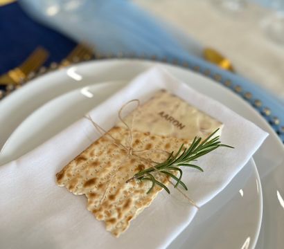 Chic place setting with stacked white plates and a folded napkin topped by a twine-tied flat cracker and fresh rosemary sprig, soft blue and gold table decor blurred in the background.