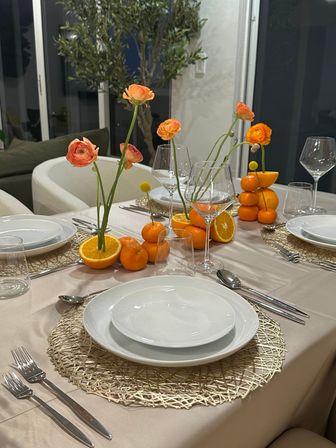 Modern dining table with white plates and wine glasses, citrus centerpiece of halved oranges and stacked mandarins holding orange ranunculus on woven placemats.