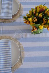 Bright spring table setting: blue-and-white striped tablecloth with white plates on fringed placemats, matching striped napkins, an empty glass, and a vase of yellow-and-red tulips centerpiece.