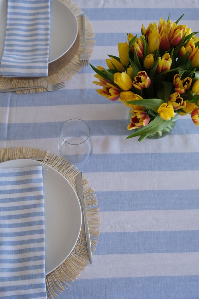 Bright spring table setting: blue-and-white striped tablecloth with white plates on fringed placemats, matching striped napkins, an empty glass, and a vase of yellow-and-red tulips centerpiece.