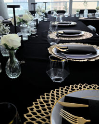 Elegant long table set with a black tablecloth, gold chargers and flatware, black napkins, clear glassware and small vases of white roses, arranged by floor-to-ceiling windows with a city skyline view.