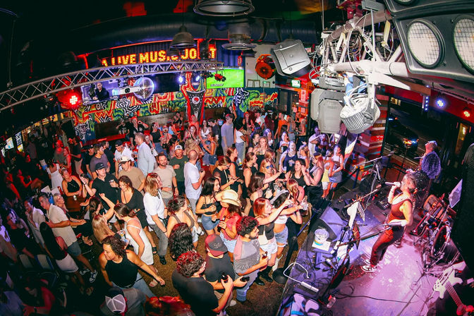 Packed live-music bar with a dancing crowd on a colorful, graffiti-lit dance floor and a band performing on stage