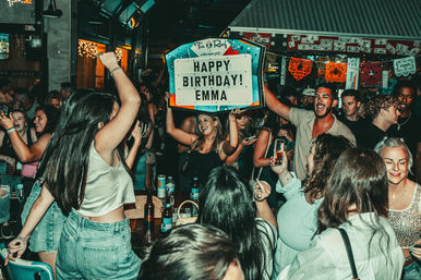 Lively bar birthday party with a crowded dance floor, friends cheering and dancing as someone holds up a lighted marquee sign reading “Happy Birthday,” drinks on tables and colorful decorations overhead.