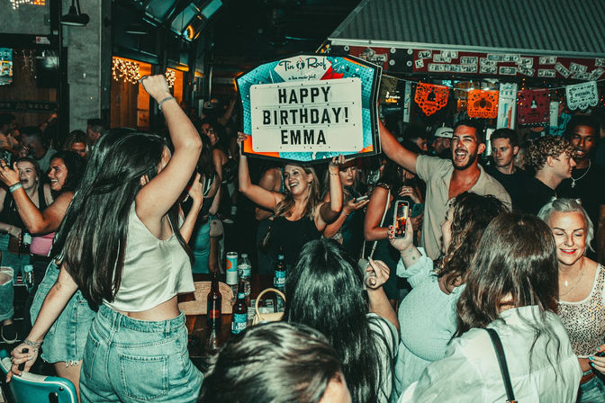 Lively bar birthday party with a crowded dance floor, friends cheering and dancing as someone holds up a lighted marquee sign reading “Happy Birthday,” drinks on tables and colorful decorations overhead.