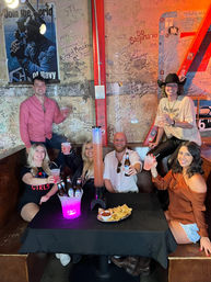 Six friends toasting in a neon-lit honky-tonk booth with graffiti-covered walls, a glowing beer bucket and tall drink tower, and chips with salsa on the table.