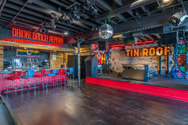 Vibrant live music bar interior with raised stage and marquee lights, disco ball over an empty dance floor, neon "Drink! Dance! Repeat!" sign, exposed brick and graffiti walls, red-and-blue bar stools at a wooden counter.