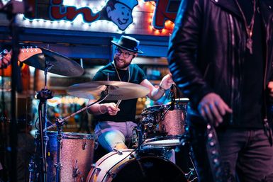 Smiling drummer in a fedora and glasses playing a sparkling drum kit on a neon-lit bar stage during a lively downtown live music night