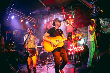 Live music at an intimate brick-walled bar stage with an acoustic guitarist, female and male vocalists, drummer, colorful stage lights and a neon script sign, nightlife performance vibe.