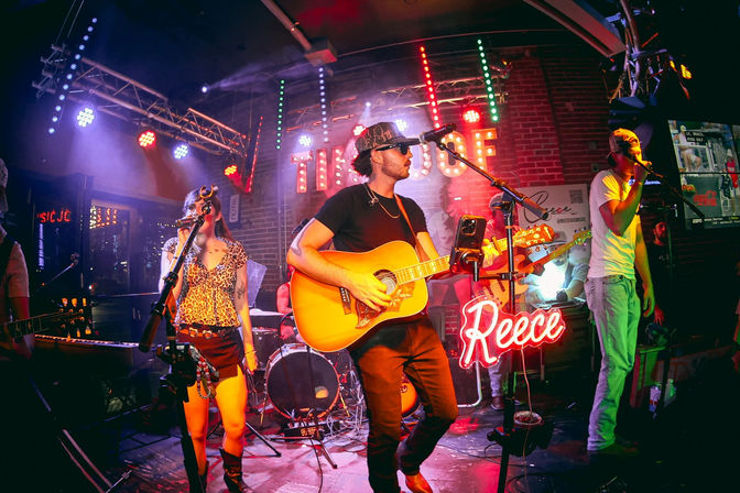 Live music at an intimate brick-walled bar stage with an acoustic guitarist, female and male vocalists, drummer, colorful stage lights and a neon script sign, nightlife performance vibe.