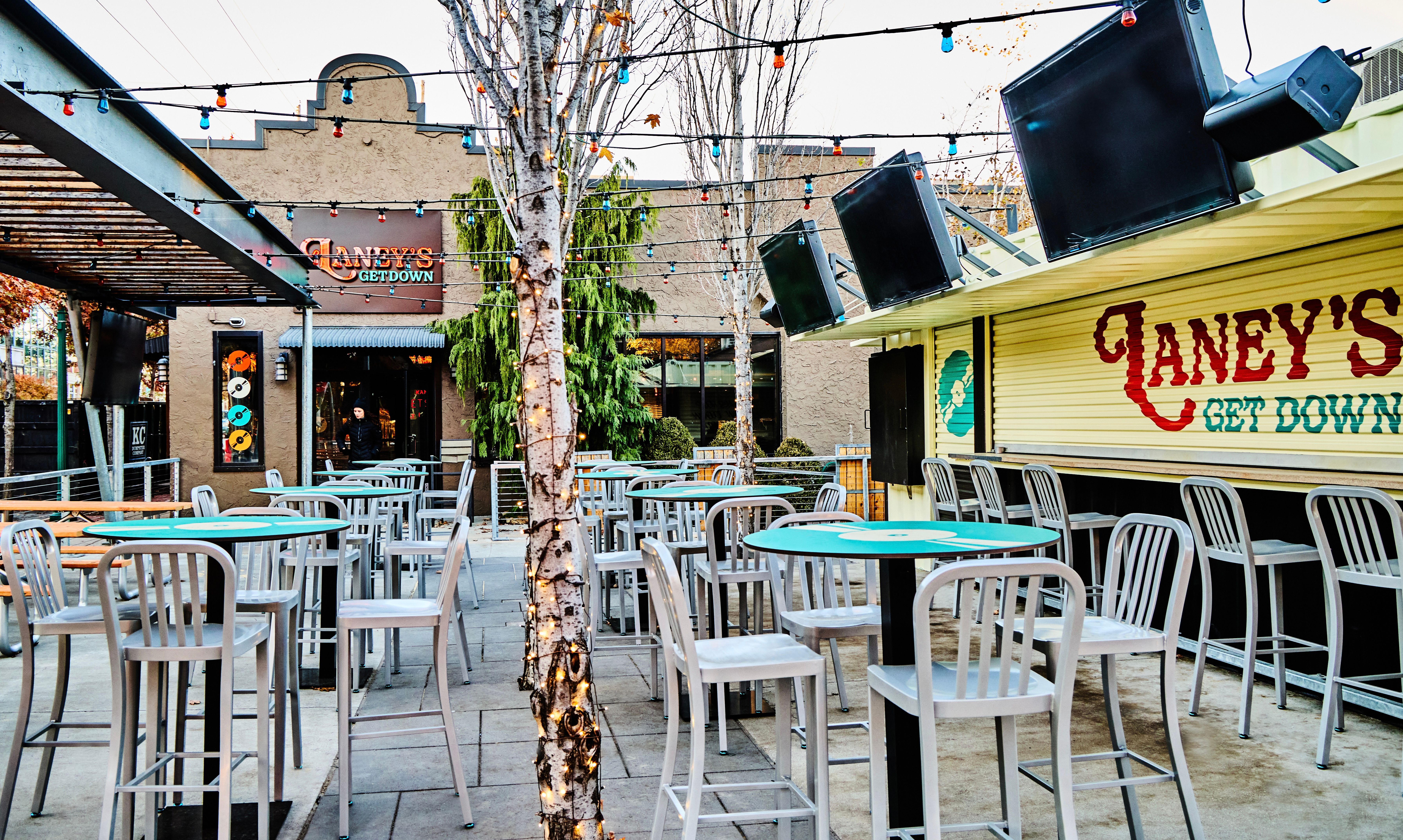Urban outdoor bar patio with string lights and lit birch trees, turquoise-topped high tables and metal stools, large wall TVs and a closed service counter with painted signage.