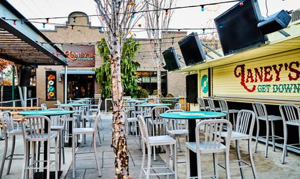 Urban outdoor bar patio with string lights and lit birch trees, turquoise-topped high tables and metal stools, large wall TVs and a closed service counter with painted signage.