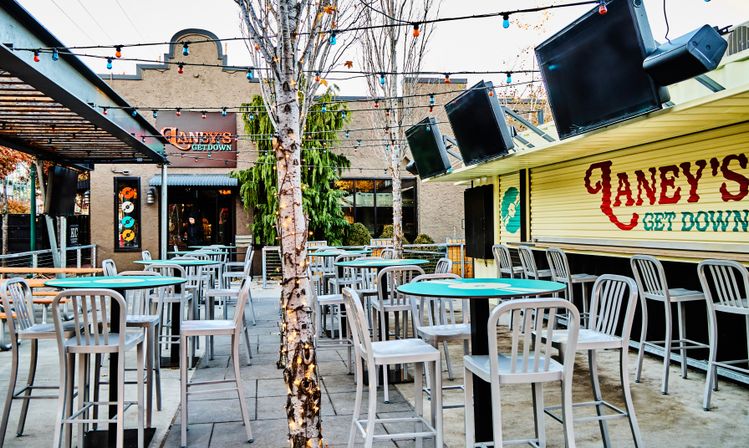 Urban outdoor bar patio with string lights and lit birch trees, turquoise-topped high tables and metal stools, large wall TVs and a closed service counter with painted signage.