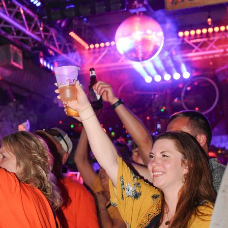 Smiling woman and party crowd raise drinks under a disco ball and vibrant purple-pink stage lights at a lively indoor nightclub